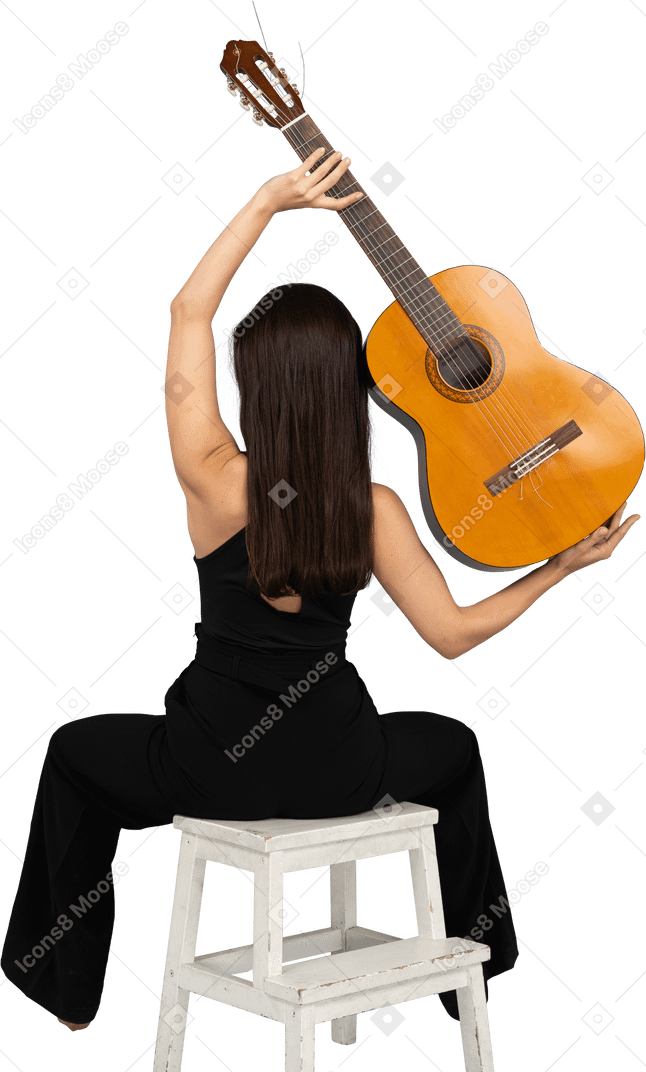 Vue arrière d'une jeune femme en costume noir tenant la guitare au-dessus de la tête et assis sur un tabouret