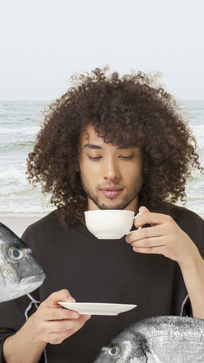 A man with curly hair drinking a cup of coffee with fish on the beach