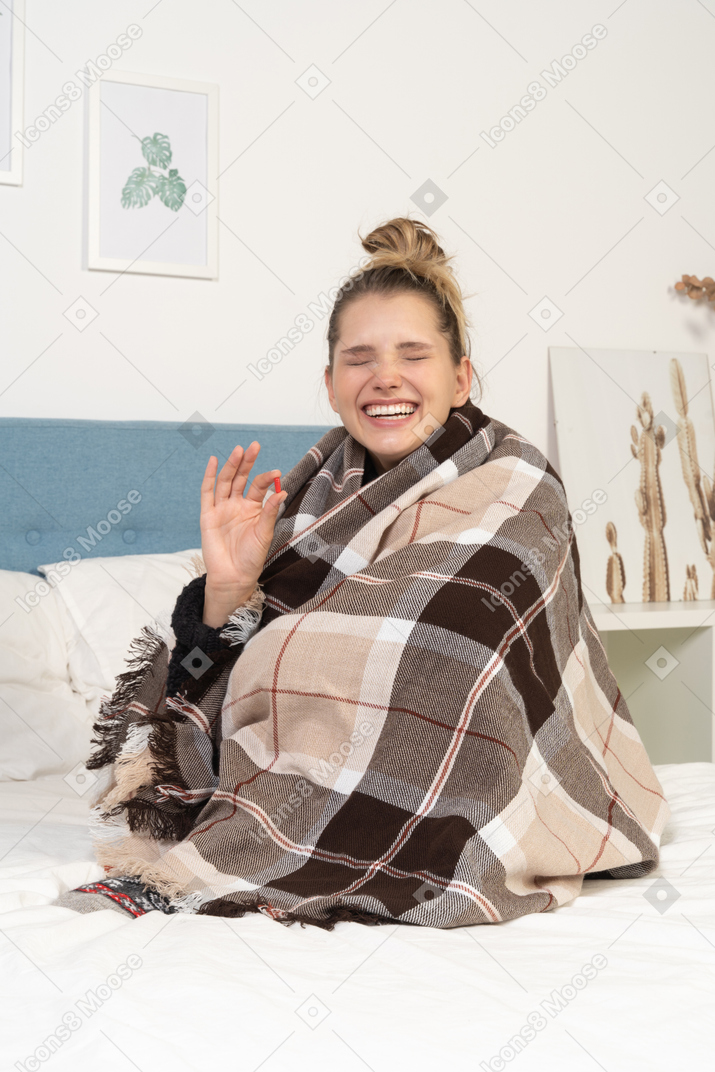 ピルを服用しているベッドでチェックの毛布に包まれて笑っている病気の若い女性の正面図