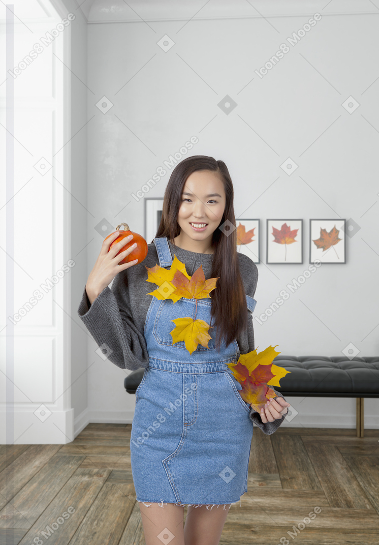 A happy young asian girl in a grey sweater and denim overalls with autumn leaves on, standing in the spacious modern-looking room with a pumpkin in her hand