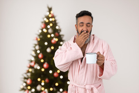 Gähnender mann mit einer tasse kaffee, der weihnachtsbaum überprüfen wird