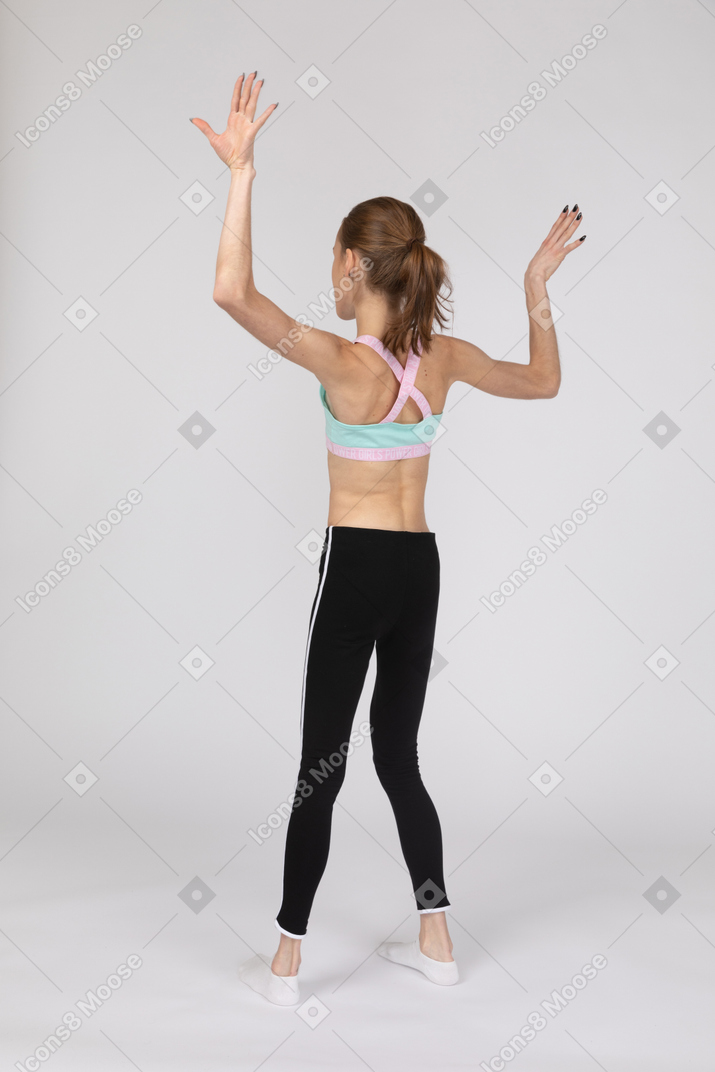 Vista traseira a três quartos de uma adolescente em roupas esportivas, levantando as mãos e dançando