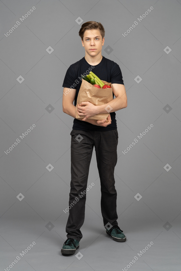 Young man standing against camera with a paper bag filled with fruit and vegetables