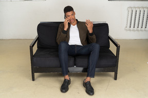 Front view of a perplexed young man sitting on a sofa and talking on his phone