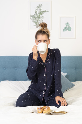 Front view of a young lady in pajamas drinking coffee in bed