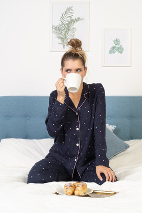 Vue de face d'une jeune femme en pyjama buvant du café au lit