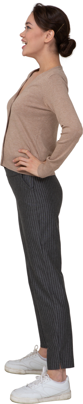 Vista laterale di una giovane donna sorridente in pullover e pantaloni mettendo le mani sui fianchi