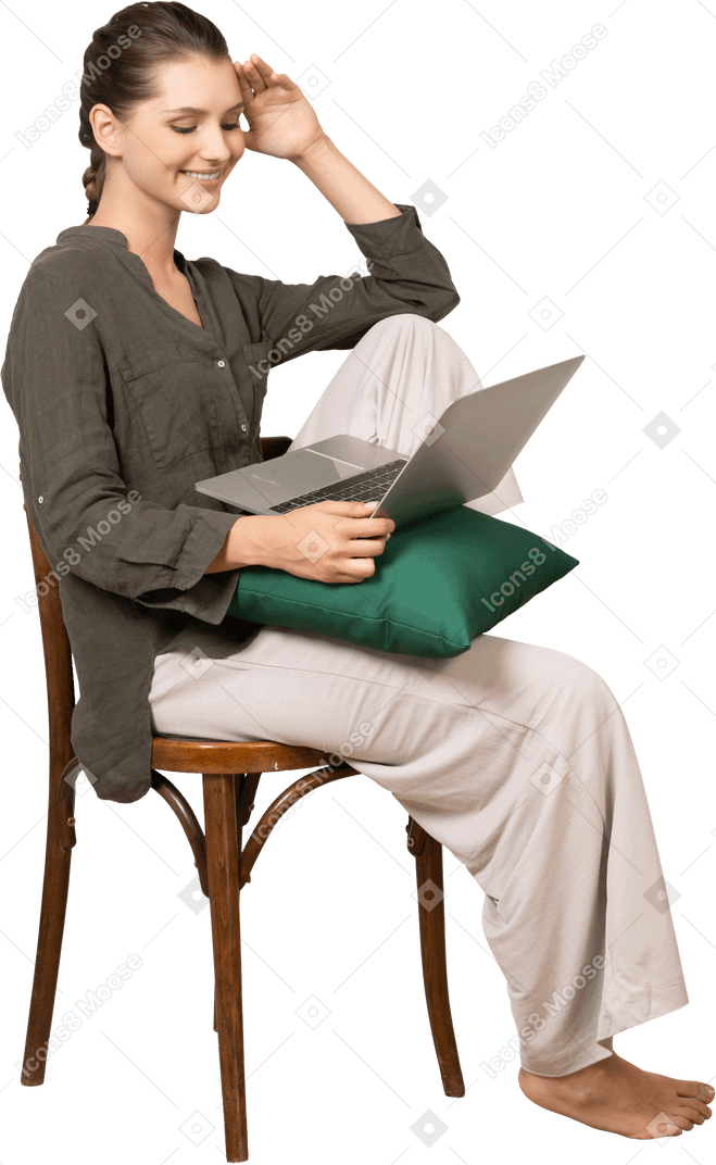 Vista lateral de una mujer joven vistiendo ropa de casa sentado en una silla con un portátil