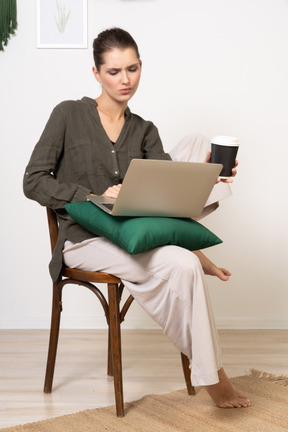 Front view of a perplexed young woman sitting on a chair and holding her laptop & coffee cup