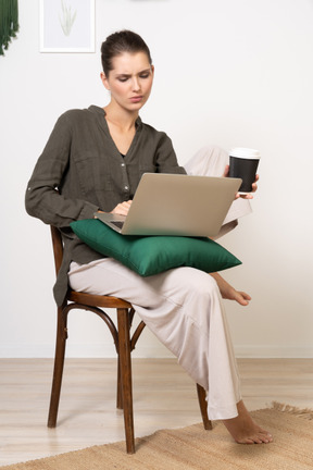 Vorderansicht einer verwirrten jungen frau, die auf einem stuhl sitzt und ihren laptop und ihre kaffeetasse hält holding