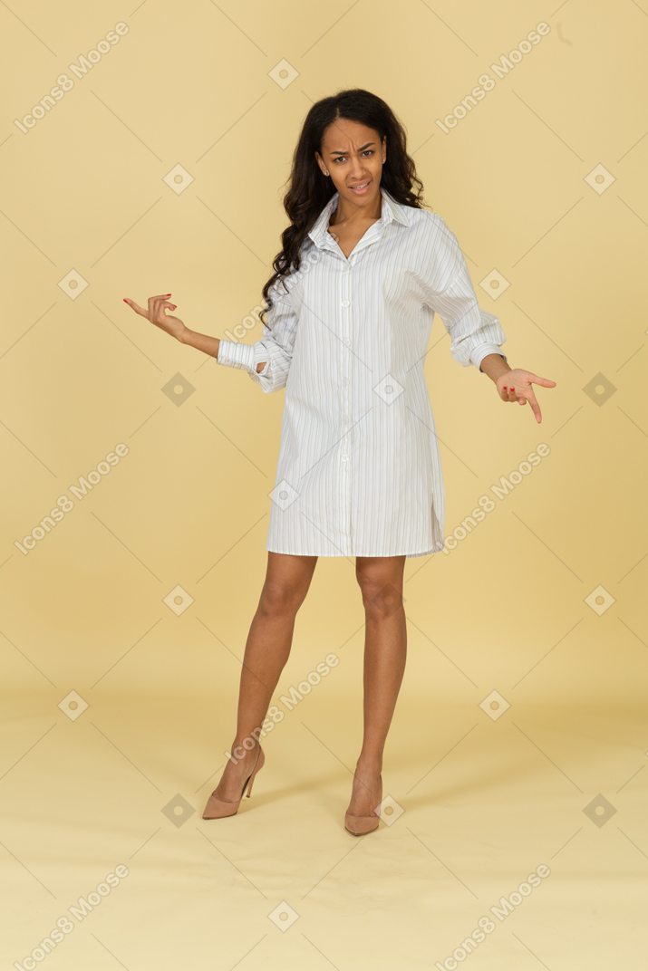 Front view of a questioning dark-skinned young female in white dress outspreading hands