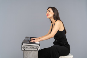 Side view of a smiling sitting young lady in black jumpsuit playing the piano