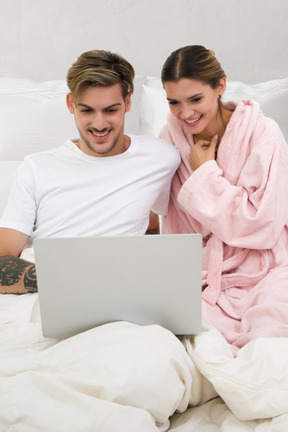 A man and woman sitting on a bed looking at a laptop