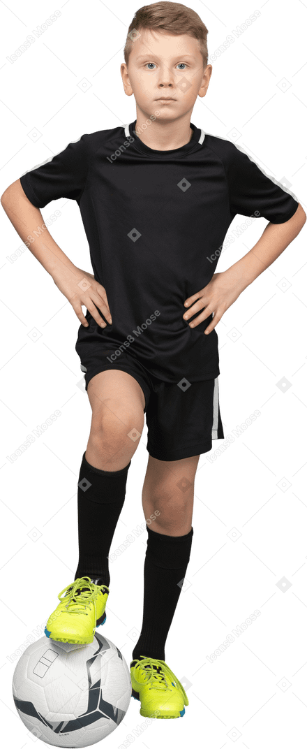 一个孩子男孩穿着橄榄球制服，将手放在臀部和他的脚在球上的前视图