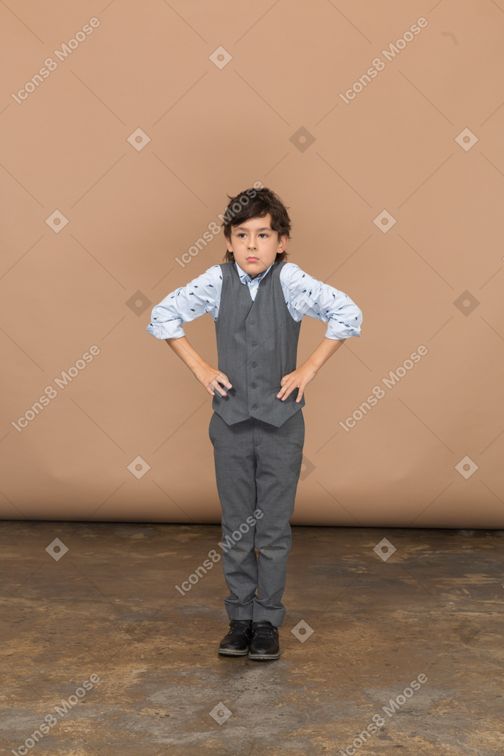 Vue de face d'un garçon mignon en costume gris posant avec les mains sur les hanches