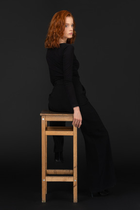 A back side view of the cute young woman sitting on the wooden chair and looking to the right