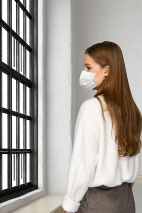 Mujer joven con mascarilla mirando por la ventana