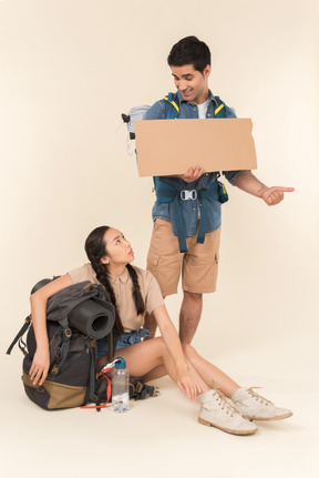 Young interracial couple showing hitchhiking sign sitting on backpacks