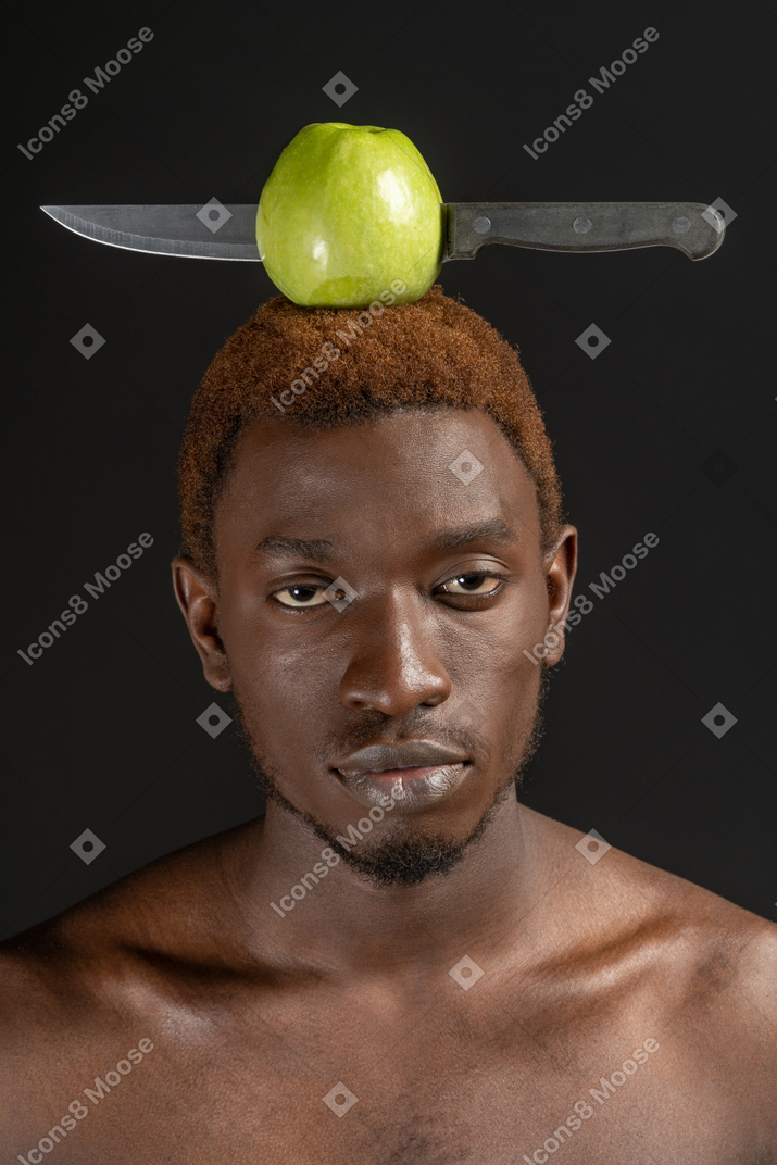 Close-up de um jovem olhando confiante para a câmera com uma maçã e uma faca na cabeça
