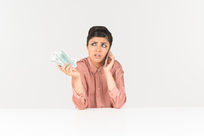 Беспокойная молодая индийская женщина держит деньги счета и разговаривает по телефону
