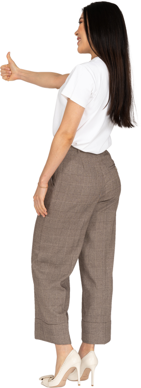 Vista posteriore di tre quarti di una giovane donna sorridente in calzoni e maglietta che mostra il pollice in su