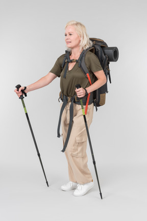 Smiling mature female tourist carrying backpack and walking with walking sticks
