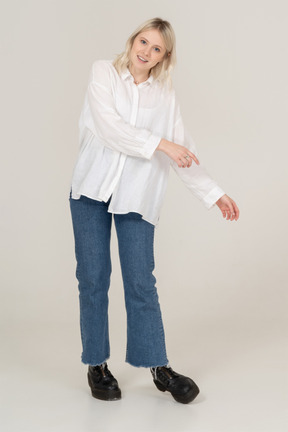 Front view of a blonde female in casual clothes stepping aside while outstretching hands and looking at camera