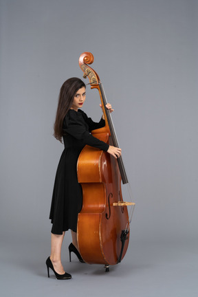 Vista lateral de un joven músico serio en vestido negro tocando su contrabajo