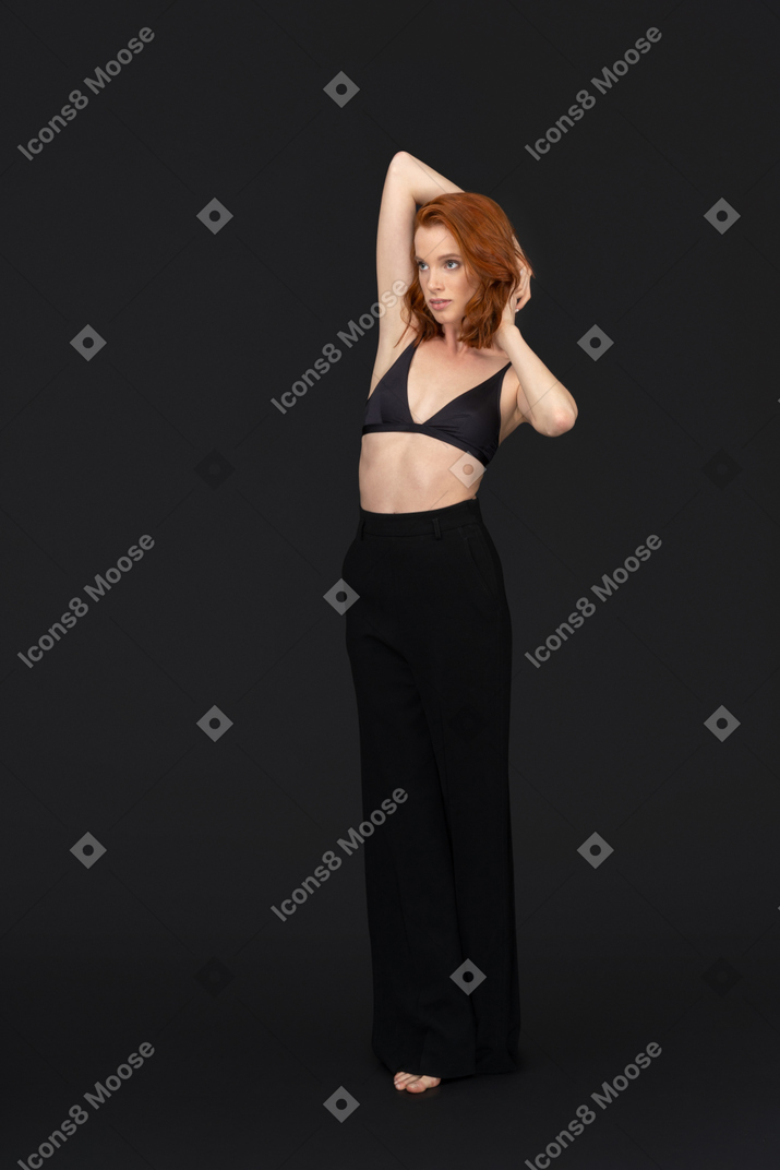 A frontal view of the sexy young woman standing on the black background an holding her hands behind the head