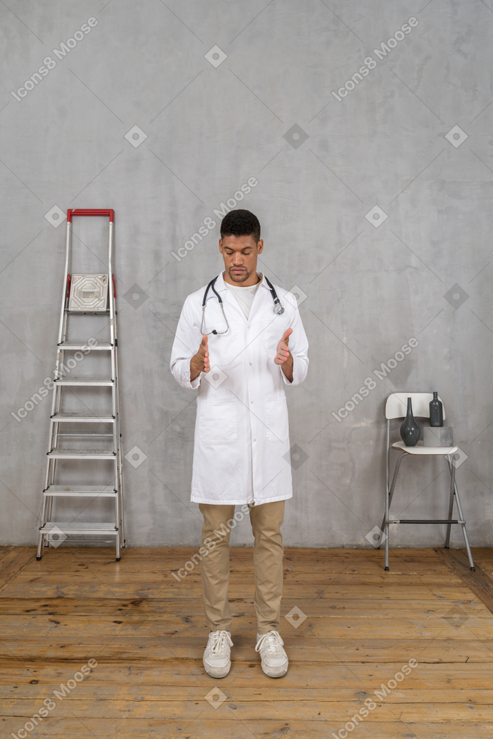 Front view of a young doctor standing in a room with ladder and chair showing a size of something