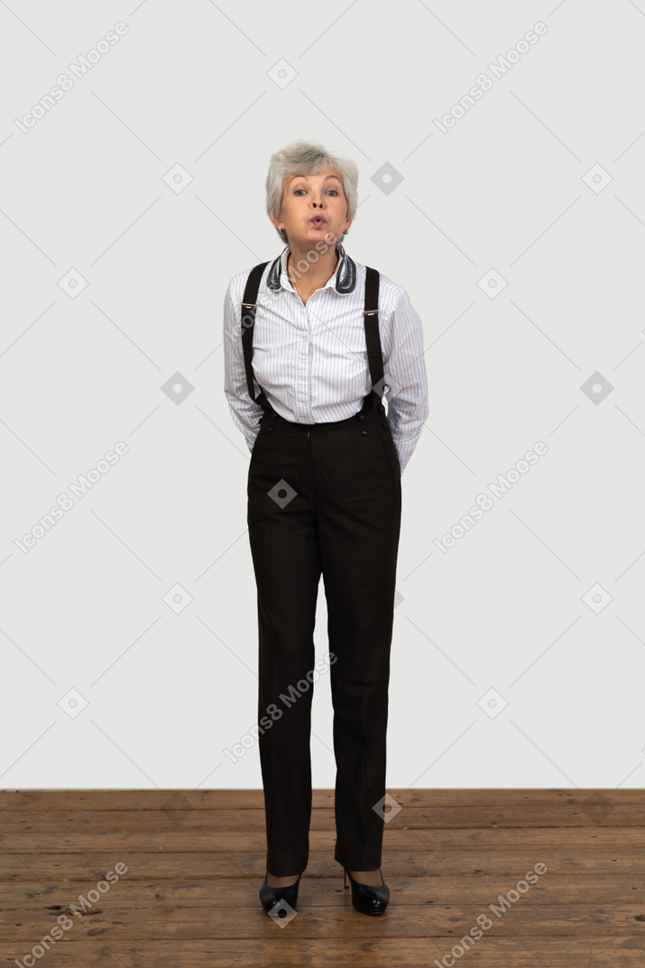 Front view of an old funny female in office clothes grimacing with her hands behind back sending a kiss