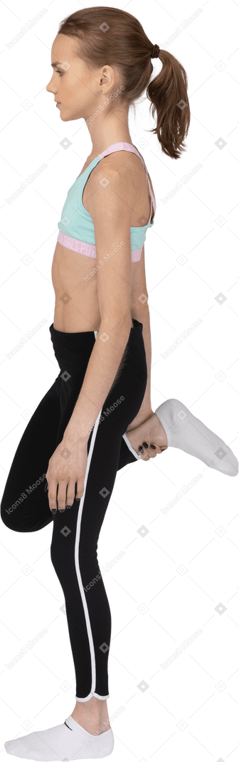 Vista lateral de una jovencita en ropa deportiva levantando la pierna y sosteniendo un tobillo
