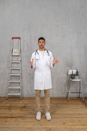 Front view of a young doctor standing in a room with ladder and chair showing a size of something