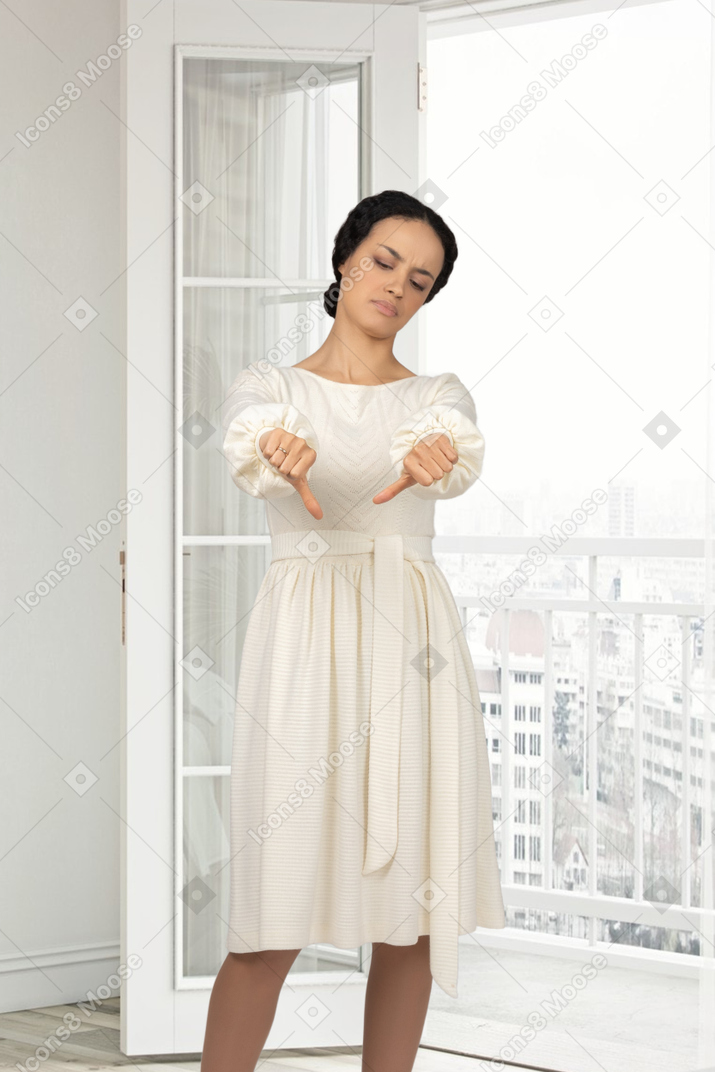 A woman in a white dress standing next to a window and showing thumbs down