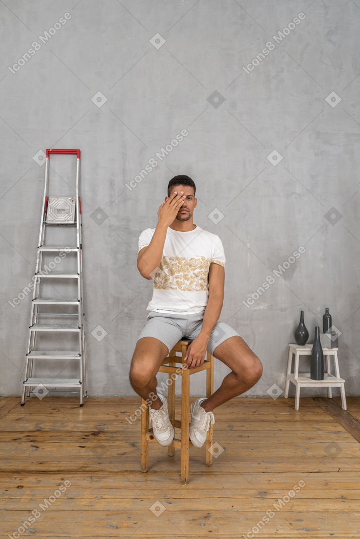 Young man sitting on stool and covering one eye
