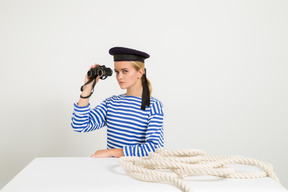 Женщина-моряк сидит за столом с веревкой на нем и держит бинокль