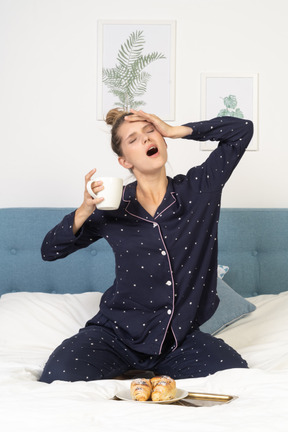 Vista frontale di una giovane donna che sbadiglia in pigiama con in mano una tazza di caffè mentre è seduta a letto