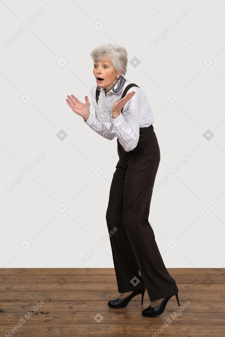 Вид в три четверти удивленной старушки в офисной одежде, поднимающей руки