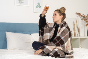 Vista laterale di una giovane donna malata in pigiama con pillole avvolte in una coperta a quadretti a letto