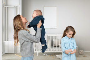 Woman holding baby up in the air next to unhappy looking daughter