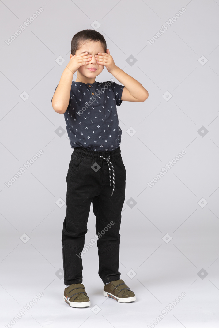 Front view of a cute boy in casual clothes covering eyes with hands