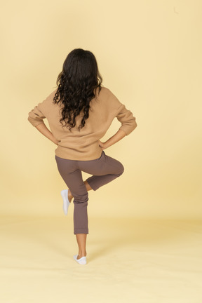 Vista posterior de una mujer joven de piel oscura levantando la pierna y poniendo las manos en las caderas