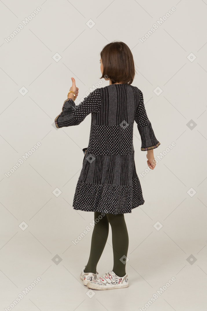 Vista traseira a três quartos de uma menina de vestido olhando para a câmera e mostrando o polegar