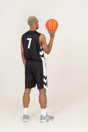 Vista posterior de tres cuartos de un joven jugador de baloncesto masculino sosteniendo una pelota