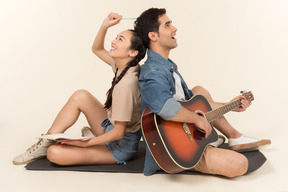 Laughing interracial couple sitting back to back and man playing guitar