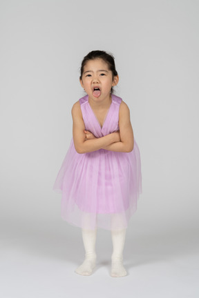 Bambina in abito rosa che tira fuori la lingua con le braccia incrociate