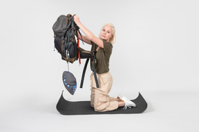 Mujer joven madura de pie sobre una alfombra turística de rodillas y levantando la mochila