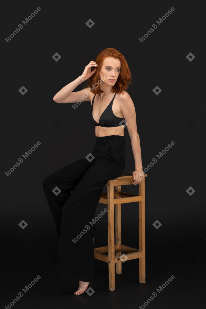 A side view of the beautiful woman dressed in black pants and bra, sitting on the wooden chair, touching her hair and looking to the right