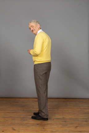 Vista traseira a três quartos de um homem idoso sorridente, vestindo um pulôver amarelo e olhando para a câmera