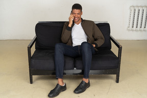 Front view of a smiling young man sitting on a sofa and talking on his phone